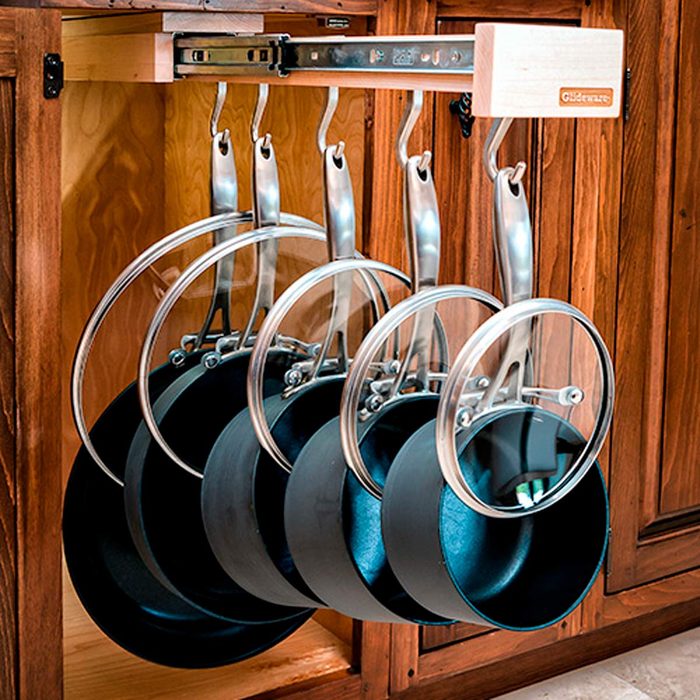 Black frying pans and glass lids hang from a metal rack inside a wooden cabinet, organized for easy access in a kitchen setting.