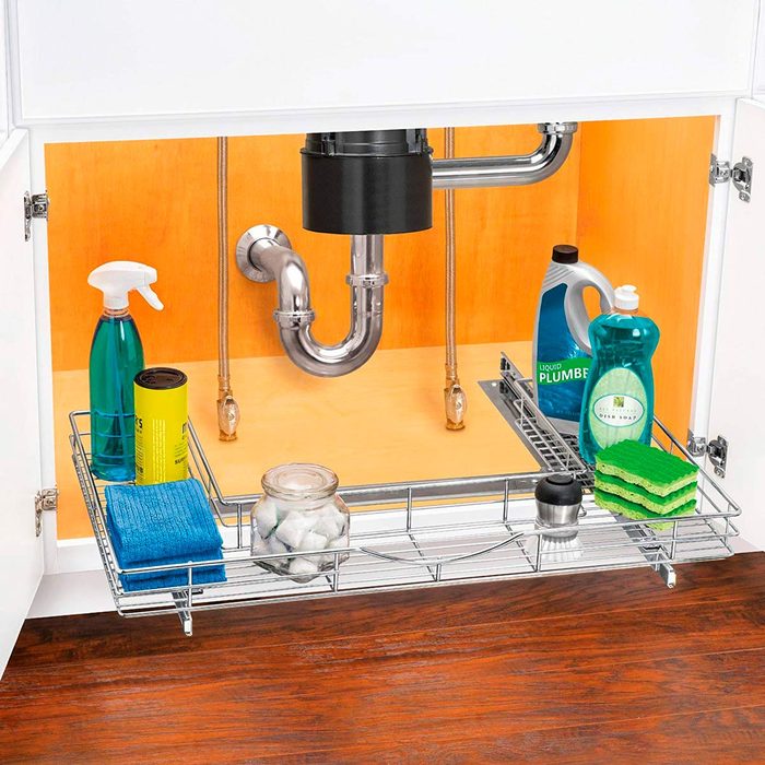 A wire shelf holds cleaning supplies like sprays and sponges under a sink, with a black disposal unit and pipes visible against a wooden backdrop.