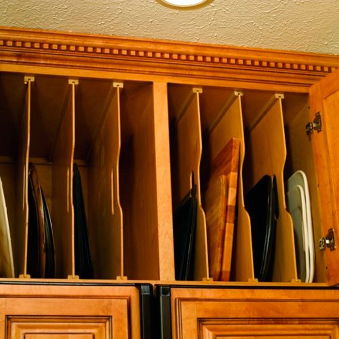 Wooden cabinet shelves hold various cutting boards and trays, neatly organized in vertical slots, illuminated by a ceiling light in a kitchen setting.