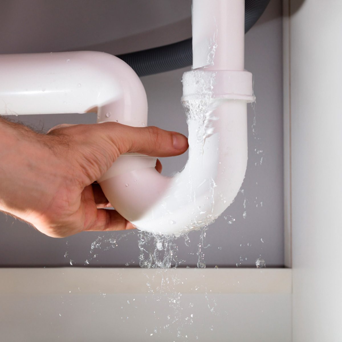 A hand grips a white plastic plumbing pipe, as water leaks from a joint, within a cabinet under a sink.