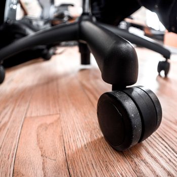 A black chair wheel rests on a wooden floor, suggesting the chair is stationary in an indoor workspace with blurred background elements.