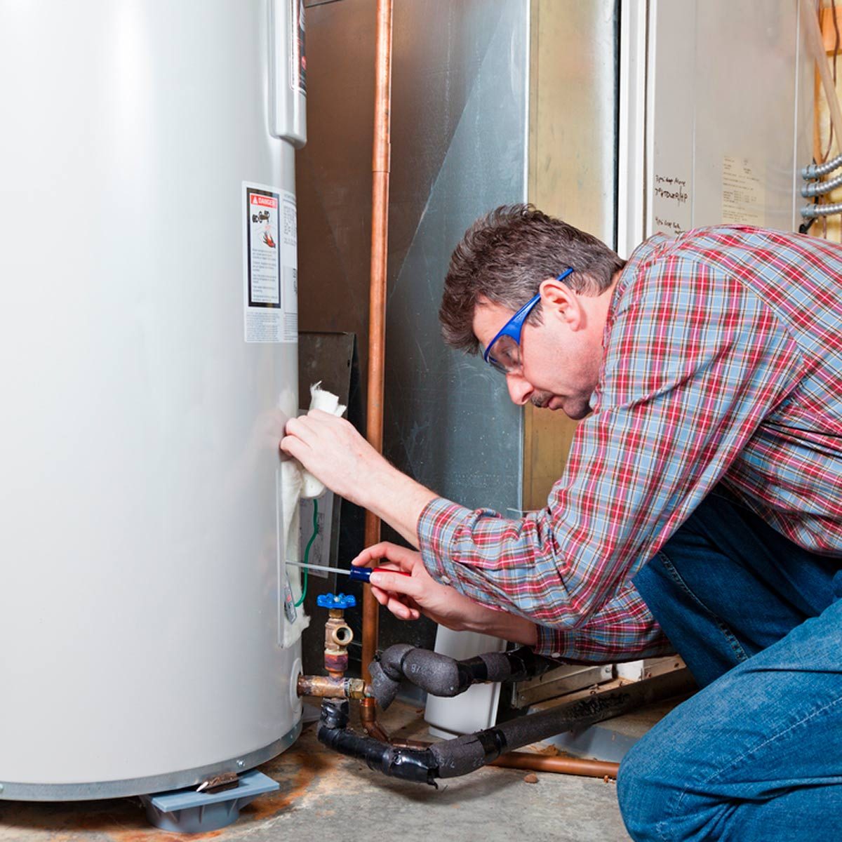 A man in protective glasses repairs a water heater, using a screwdriver on a valve while crouched on a concrete floor in a utility area.