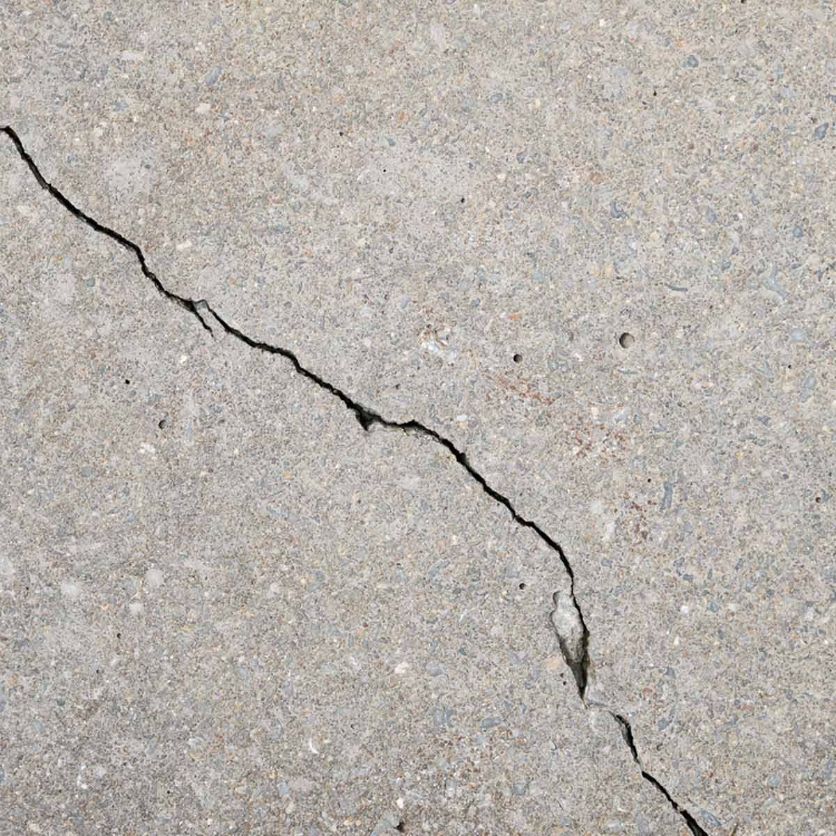 A cracked concrete surface shows an irregular fissure extending diagonally, revealing a textured gray material with small stones embedded throughout.