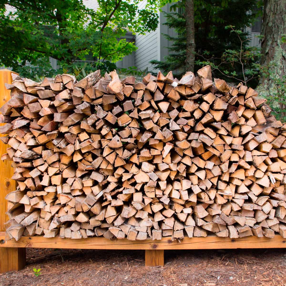 A neatly stacked pile of firewood rests on a wooden rack, surrounded by greenery and trees, indicating an outdoor setting.