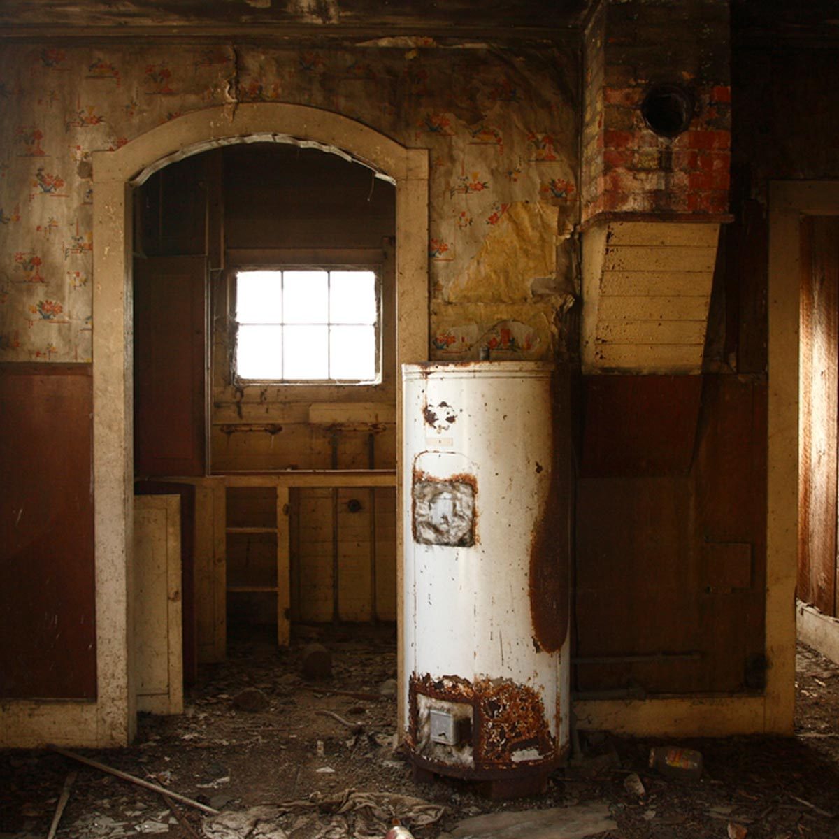 A rusty water heater stands in a dilapidated room, surrounded by peeling wallpaper and debris, with a small window providing faint light.