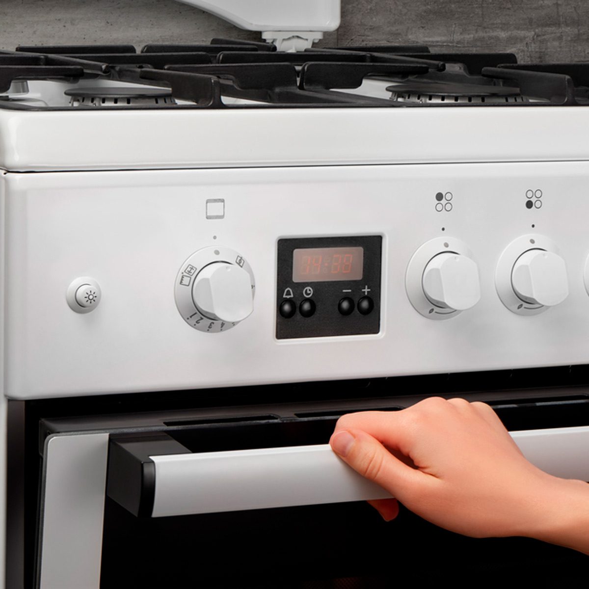 A hand is pulling open a white oven door, revealing control knobs and a digital timer, set against a textured gray wall background.