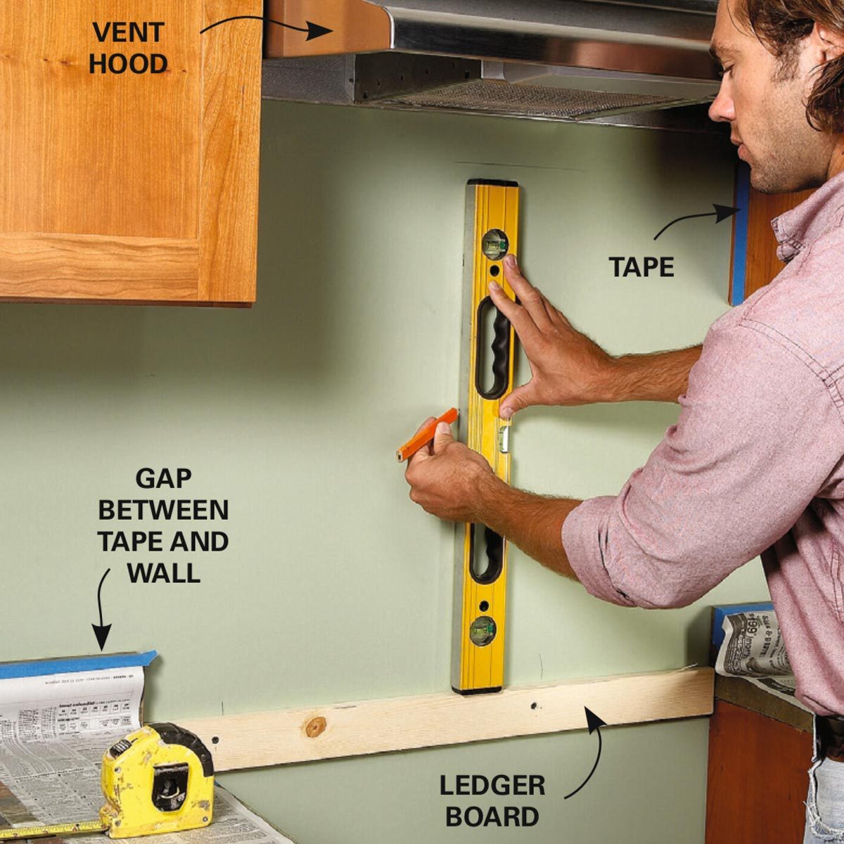 A man uses a level tool against a wall while marking measurements for a ledger board installation, with cabinetry and a vent hood above.