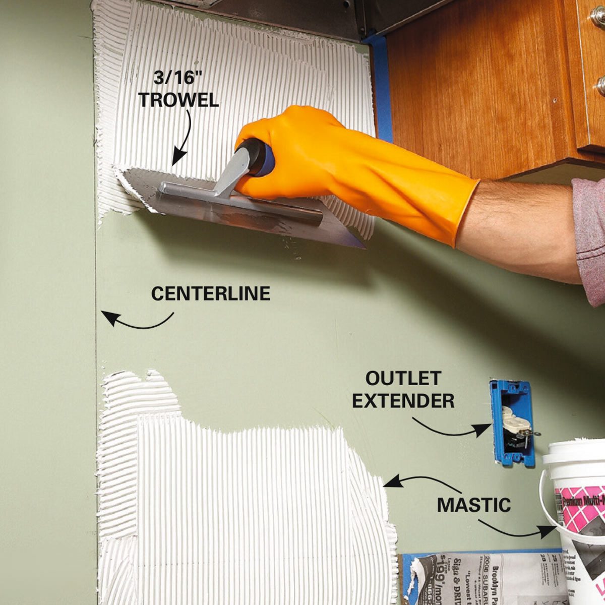 A hand with an orange glove holds a trowel, spreading adhesive on a green wall, near an electrical outlet and a bucket of mastic.