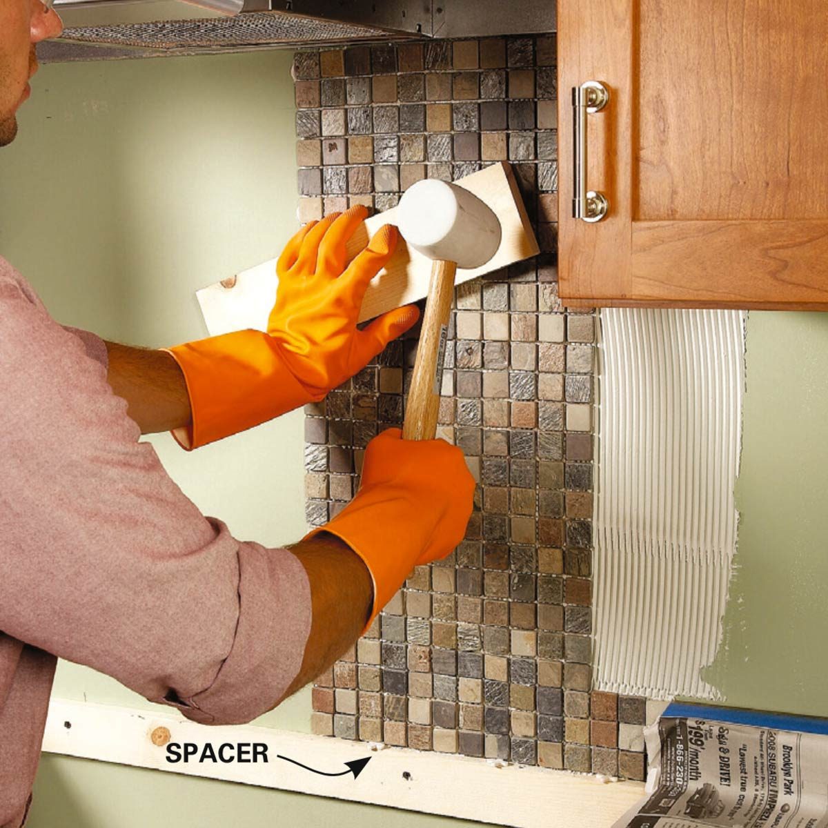 A person wearing orange gloves uses a mallet to tap a wooden board against a tiled wall during a kitchen remodeling project.
