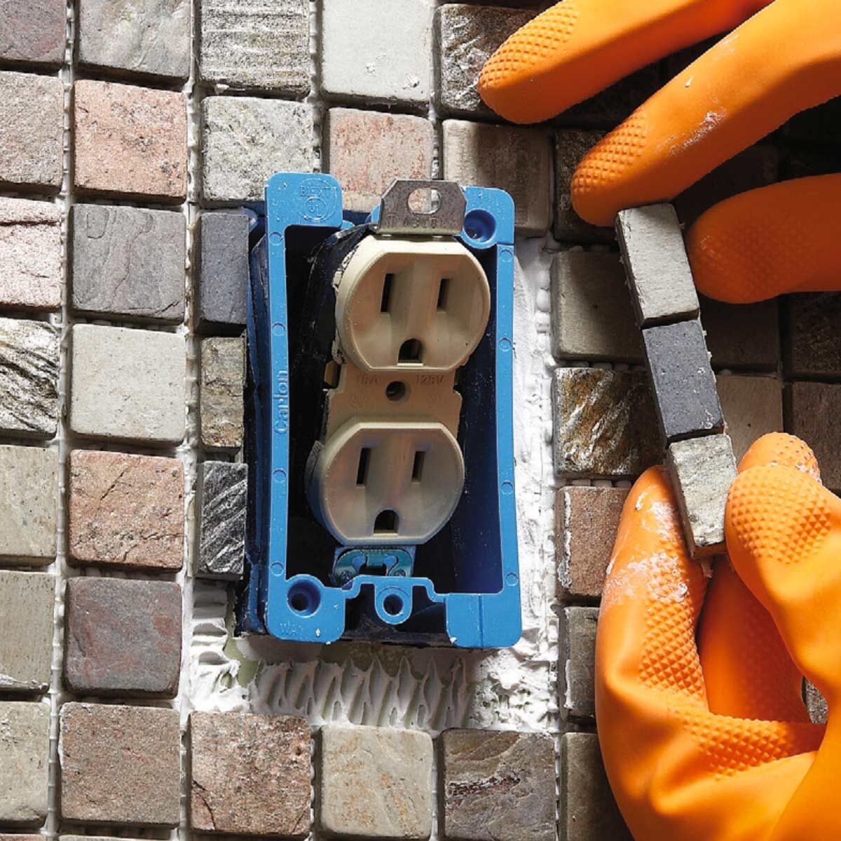 A power outlet is mounted in a wall, while a gloved hand positions a tile piece nearby among various stone tiles.