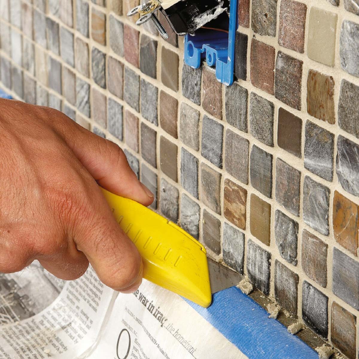 A hand using a yellow utility knife is scraping grout off a tiled wall near a blue electrical box, with newspaper laid beneath for protection.
