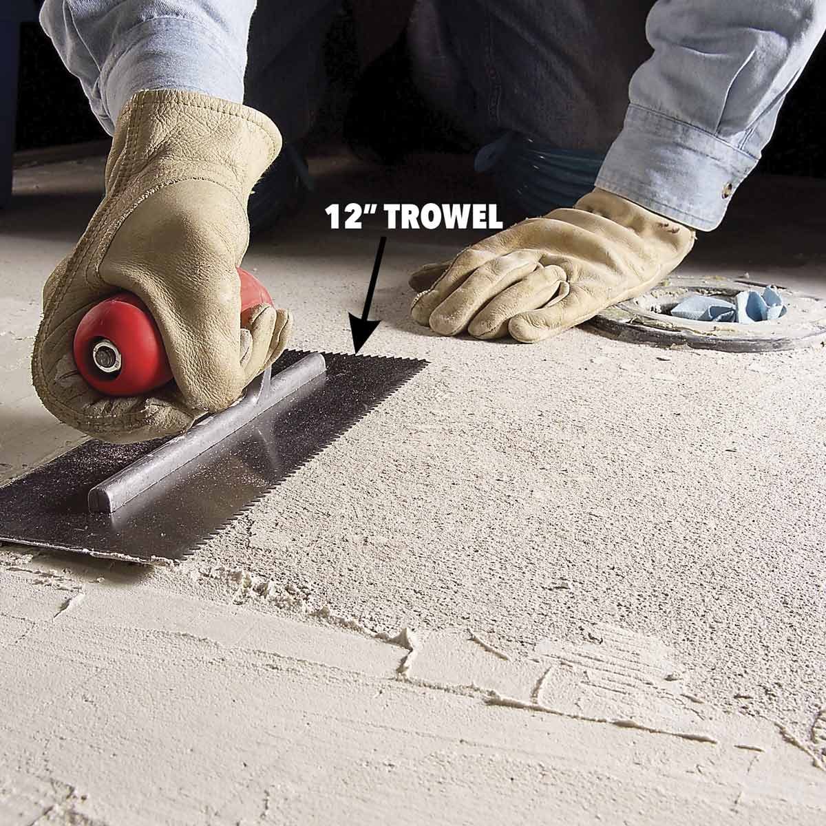 A gloved hand holds a 12-inch trowel, smoothing a textured surface on the floor, while another gloved hand rests nearby.