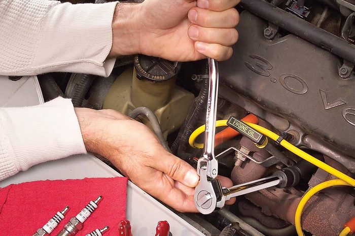 A hand uses a ratchet wrench to tighten a spark plug in a car engine, surrounded by automotive tools and spark plugs on a red cloth.
