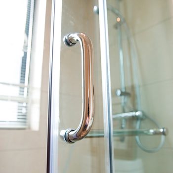 A polished metal handle is mounted on a glass shower door, providing grip. Soft light filters through a nearby window, illuminating tiled walls.