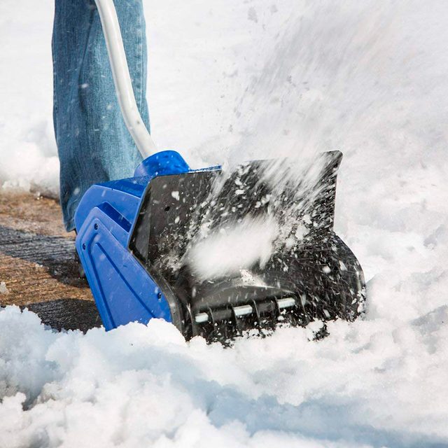 A blue snow shovel pushes snow aside, spraying flakes into the air, on a concrete surface surrounded by a snowy landscape.