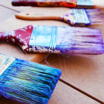 Multiple paintbrushes with colorful bristles are laid out on a wooden surface, showcasing various shades of blue and purple, some with remnants of paint.