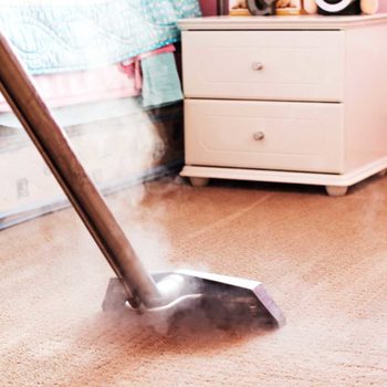 A vacuum cleaner is actively cleaning a carpet, releasing steam, in a bedroom with a bed and nightstand visible in the background.