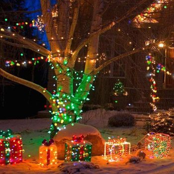 Colorful, illuminated presents are arranged under a tree decorated with lights, set in a snowy yard at night. Vibrant holiday ambiance surrounds the scene.