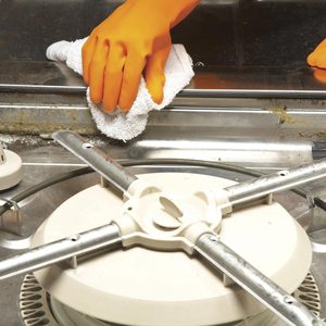 A gloved hand wipes a metal surface with a white cloth, cleaning away dirt in a kitchen environment around a dishwashing appliance.