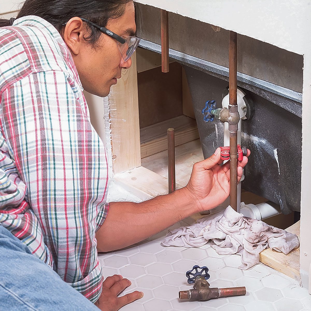 A person adjusts a plumbing valve under a sink, surrounded by pipes, a wrench, and a cloth on the white tiled floor.