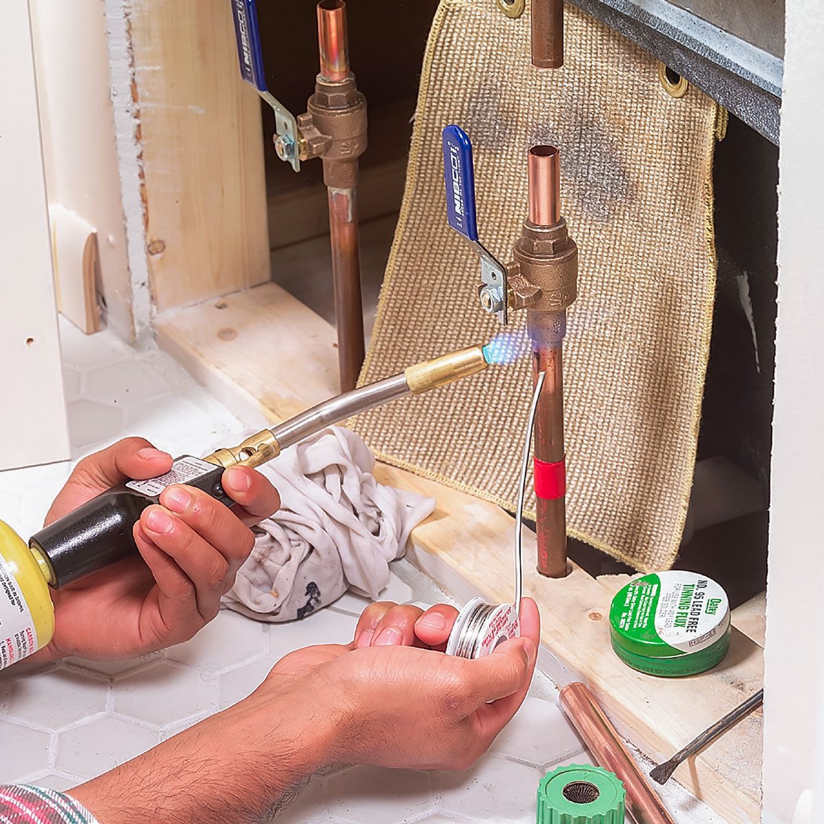 A person uses a blowtorch to solder copper pipes, surrounded by plumbing tools and materials on a tiled floor inside a cabinet.