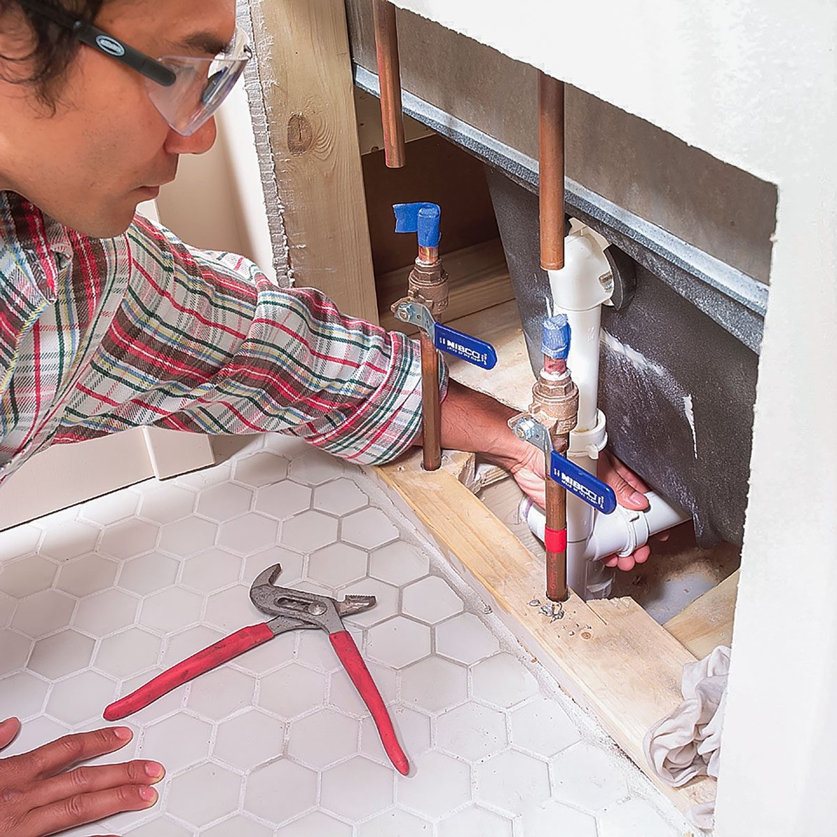 A man is crouching under a sink, using his hand to manipulate plumbing pipes while a pair of red-handled pliers rests on the tiled floor nearby.