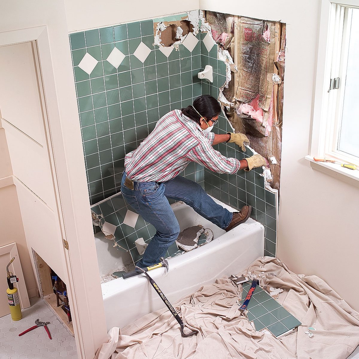 A person is removing broken tiles from a bathroom wall, standing in a bathtub. Surrounding debris is on a drop cloth, and tools are nearby.