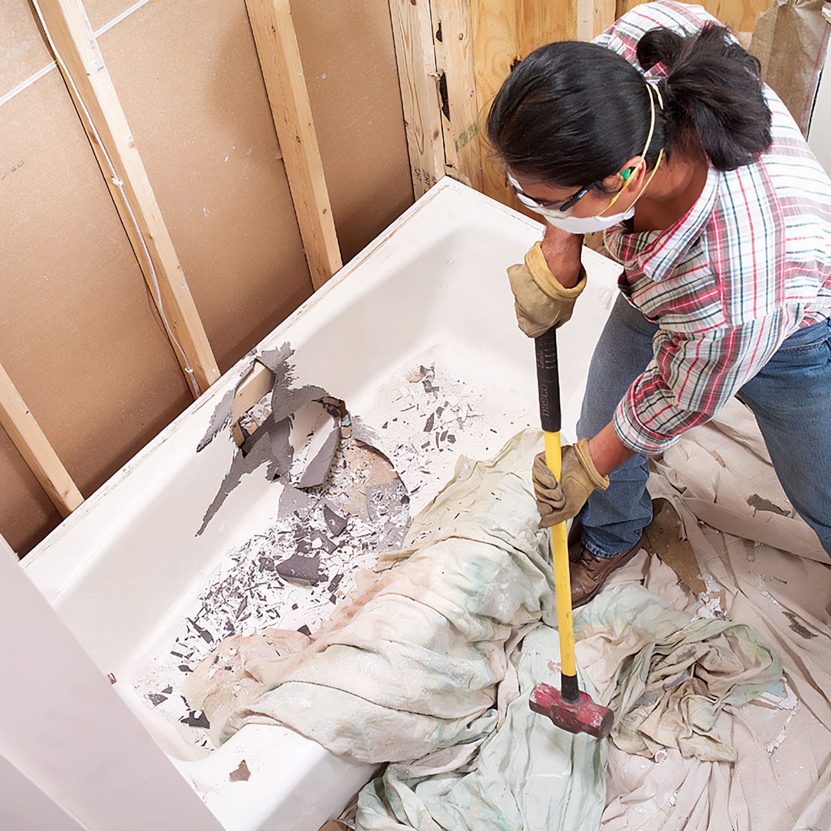 A person in gloves removes debris from a bathtub using a sledgehammer, surrounded by a partially constructed wall and protective coverings on the floor.