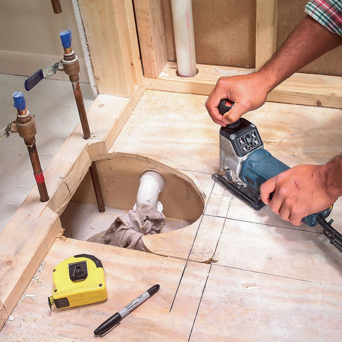 A person uses a jigsaw to cut a hole in a wooden floor near plumbing pipes, surrounded by tools like a tape measure and marker.