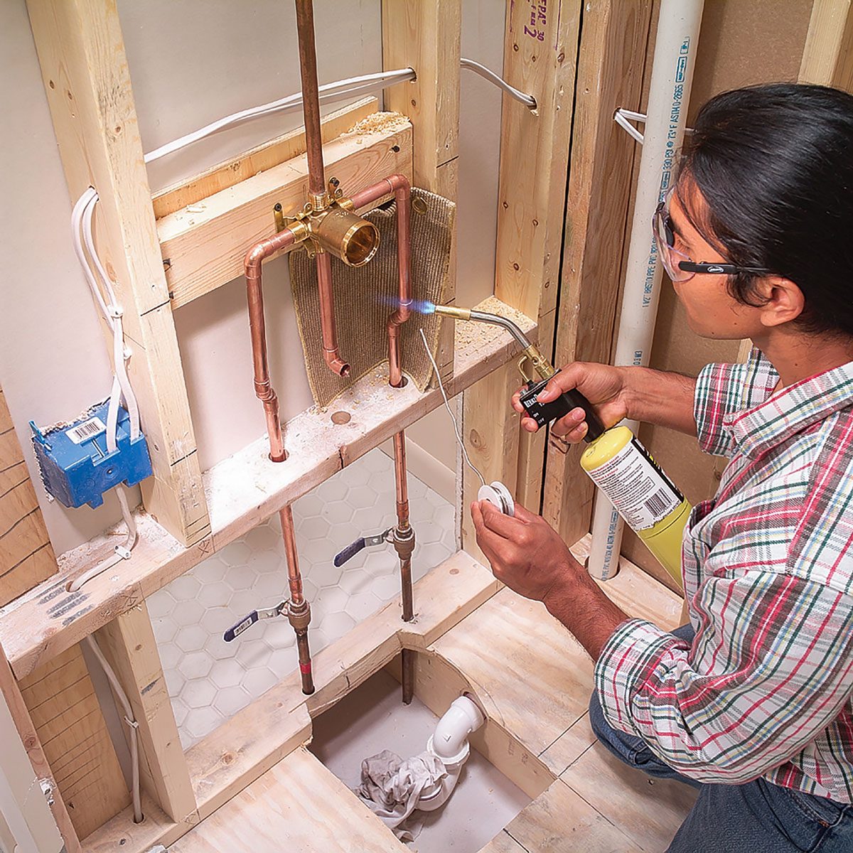 A person uses a torch to solder copper pipes in a bathroom under construction, surrounded by wooden framing, plumbing fixtures, and expose walls.