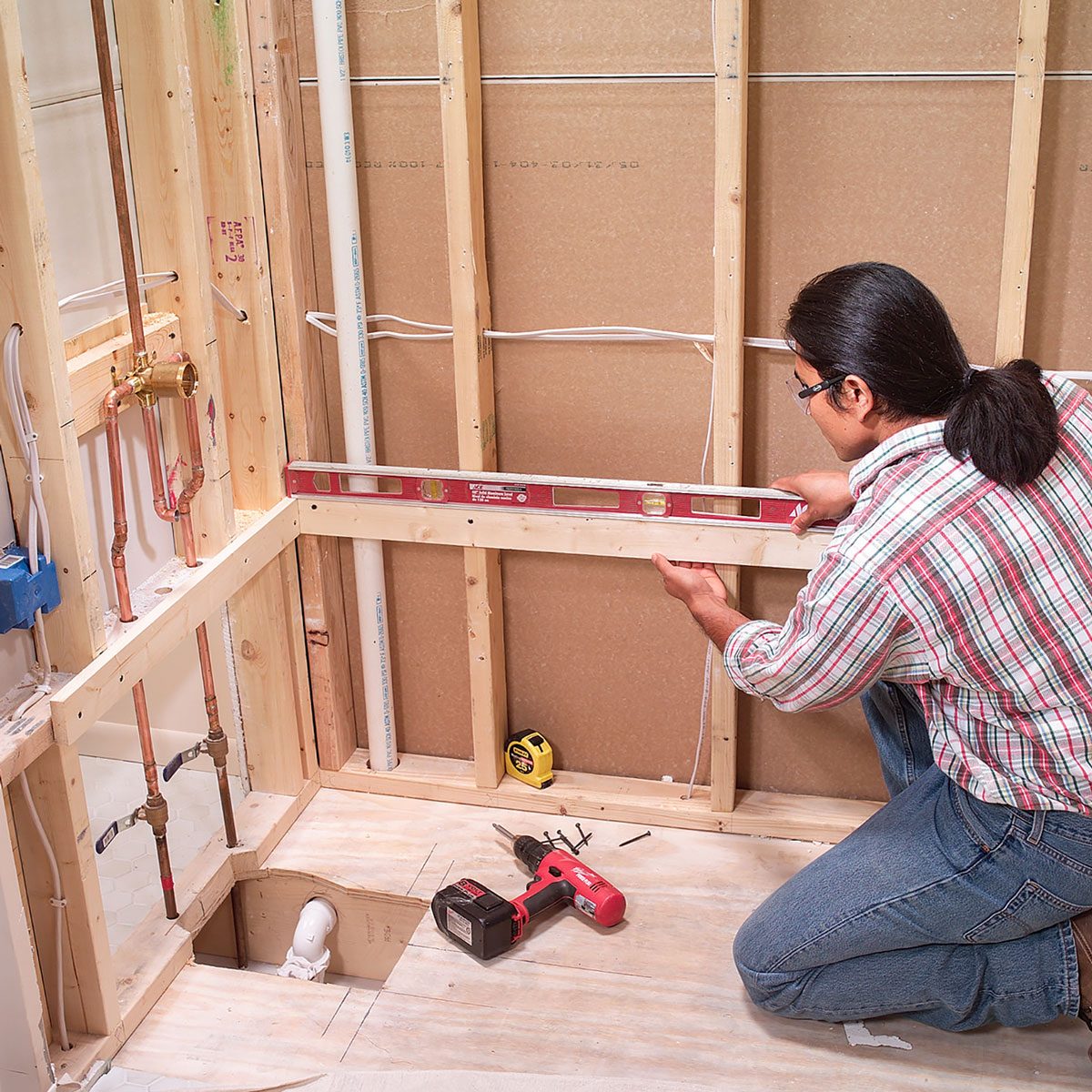A person uses a level on wooden framing in a construction area, surrounded by plumbing pipes and tools, including a drill and measuring tape.