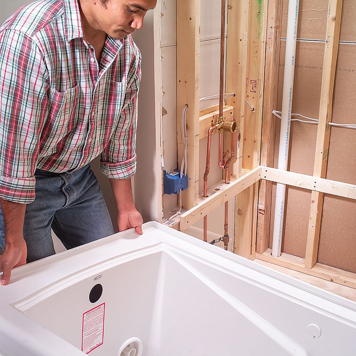A person lifts a white bathtub into place within a partially constructed bathroom, surrounded by exposed wooden framing and plumbing fixtures.