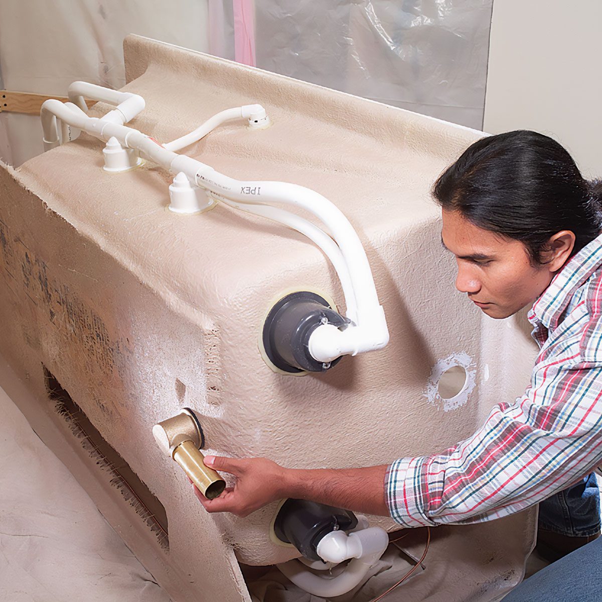 A person installs plumbing components on a large, beige fixture while positioned against a light-colored background, focused on connecting pipes and fittings.