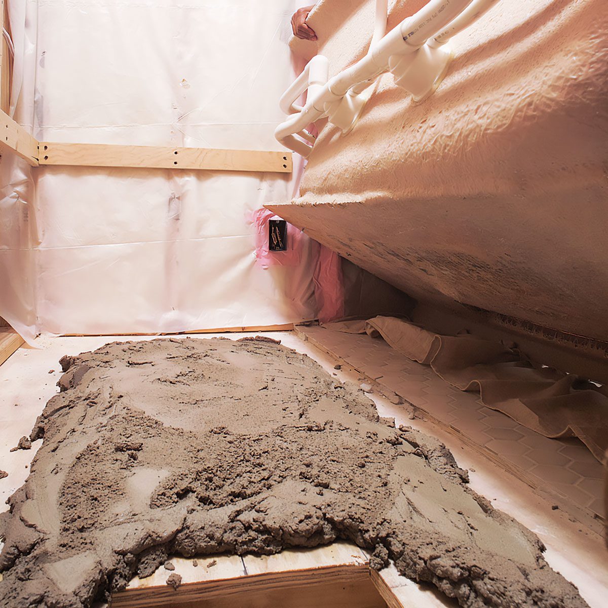 A patch of wet concrete is spread across a wooden floor inside a partially enclosed space, with walls covered in plastic sheeting and a sloped ceiling.
