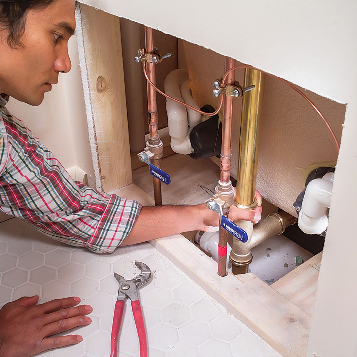 A person uses a wrench to tighten plumbing connections beneath a sink, surrounded by copper and plastic pipes in a domestic environment.