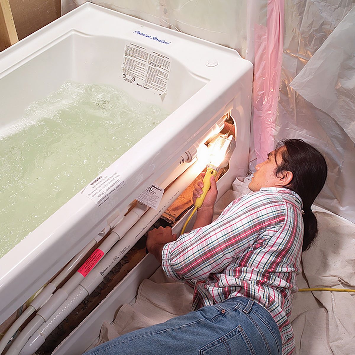 A person lies next to a hot tub, holding a flashlight while working on its plumbing, surrounded by plastic sheeting and a tarp-covered floor.