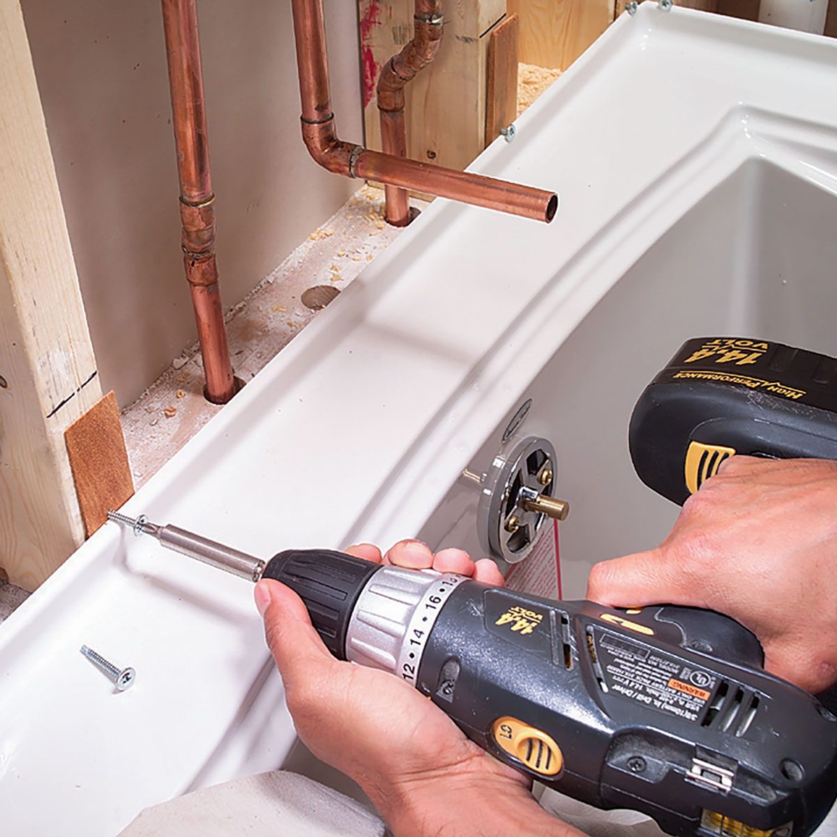 A person uses a cordless drill to fasten a screw into a bathtub, surrounded by plumbing pipes and construction materials in a partially completed bathroom.