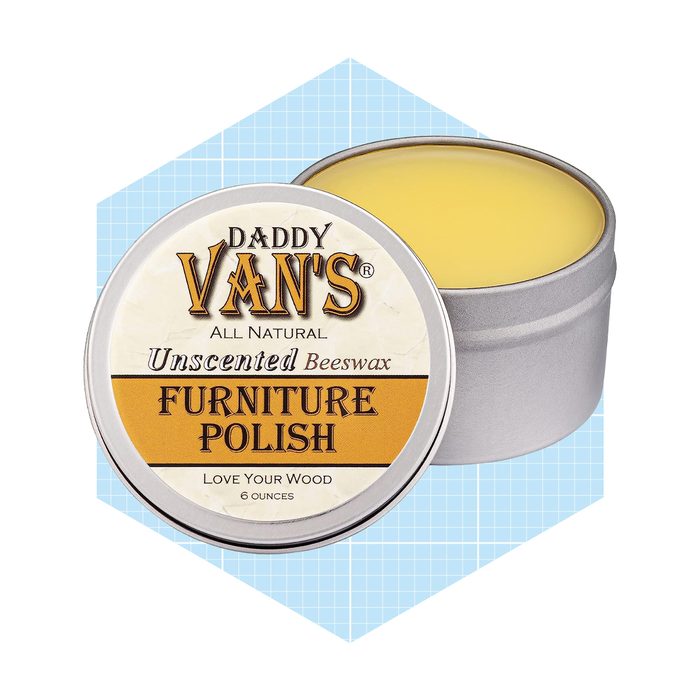 A round tin of unscented beeswax furniture polish is shown, half-open, against a light blue geometric background. The label reads 