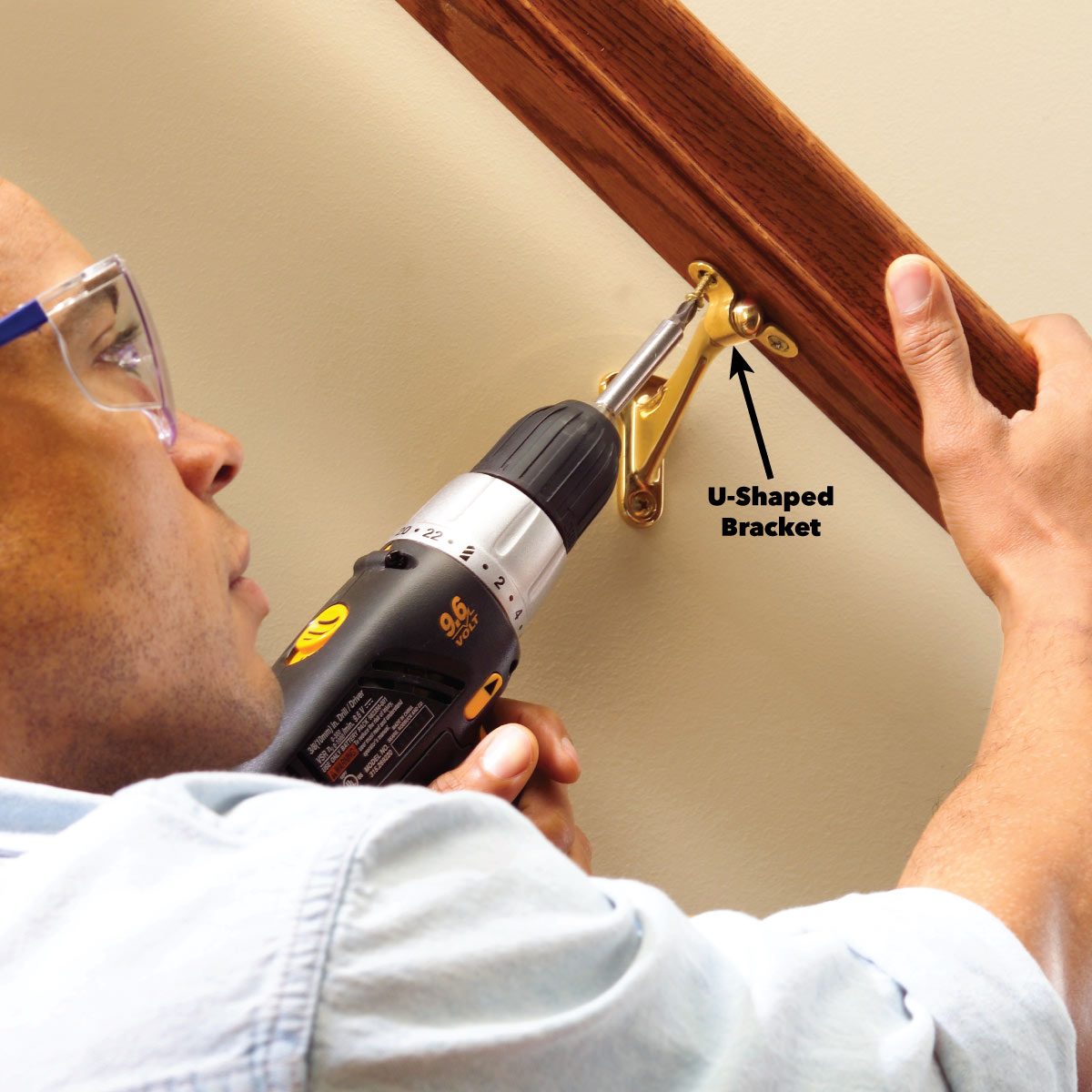 A person is using a power drill to secure a U-shaped bracket to a wall, which supports a wooden railing above it.