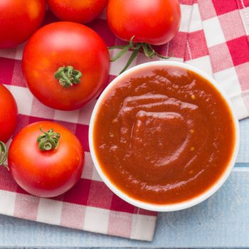 A bowl of red tomato sauce sits near fresh tomatoes on a red and white checkered cloth, resting on a blue wooden surface.