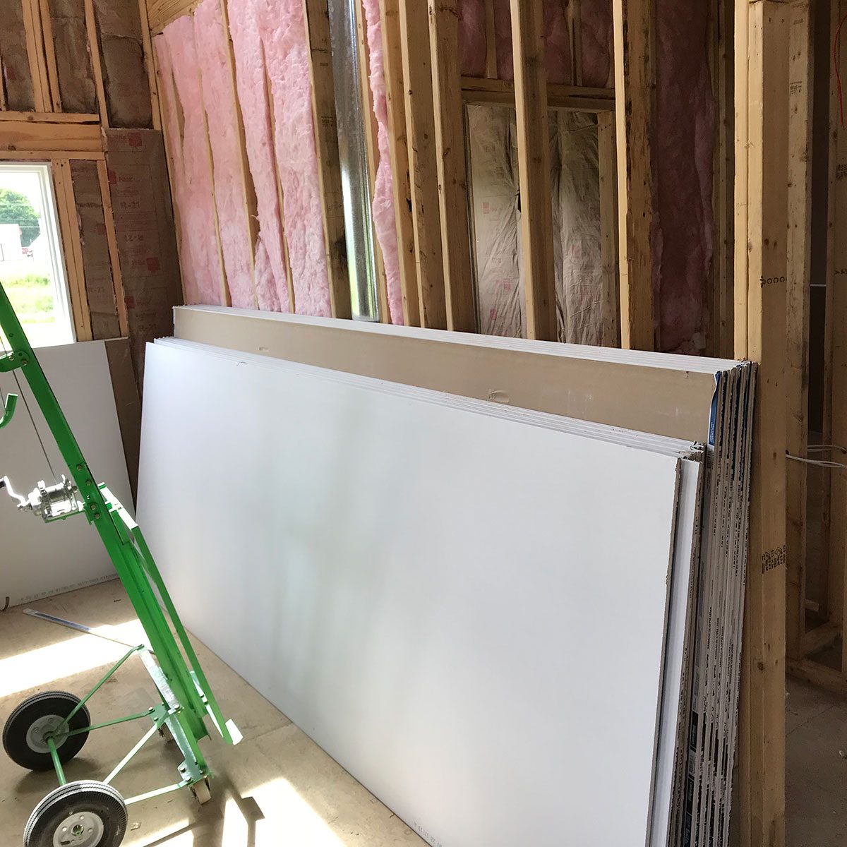 Stacks of drywall panels lean against a wall, while a green dolly sits nearby in a partially constructed room with exposed wooden framing and insulation.