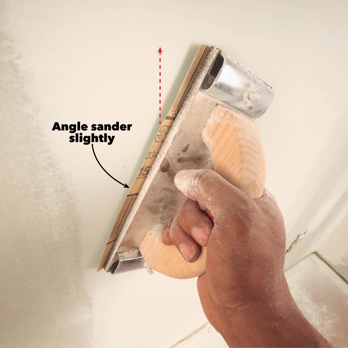 A hand holds an angle sander, smoothing a wall. Powder dust is visible, indicating recent smoothing activity in a light-colored, unfinished indoor space.