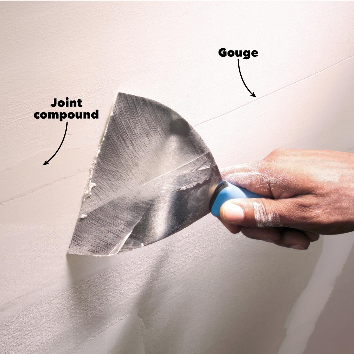 A hand holds a metal putty knife, smoothing joint compound on a wall, removing imperfections in a light-colored, unfinished drywall surface.