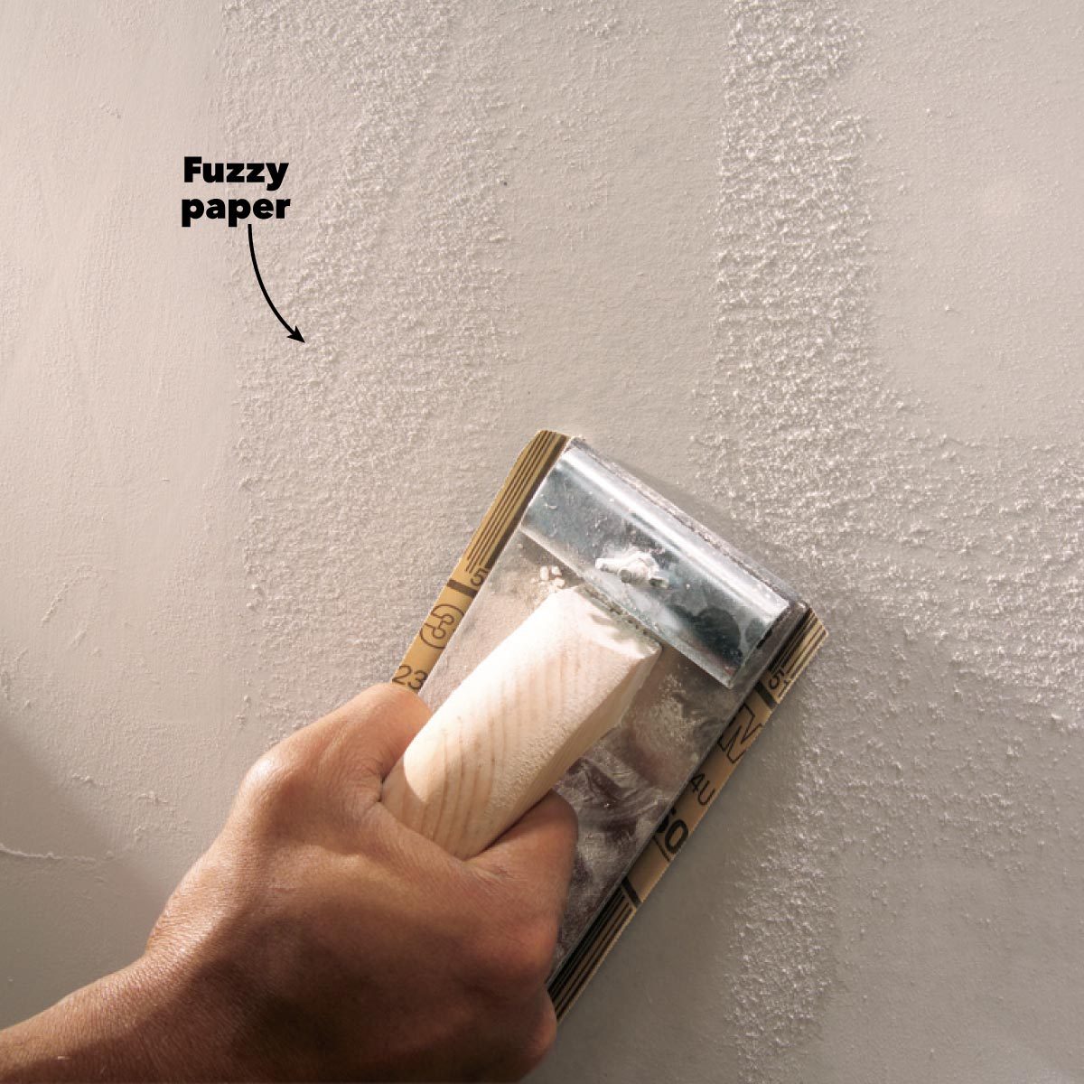A person is sanding a textured wall with a hand-held tool, smoothing out uneven areas in a light-colored, unfinished surface.