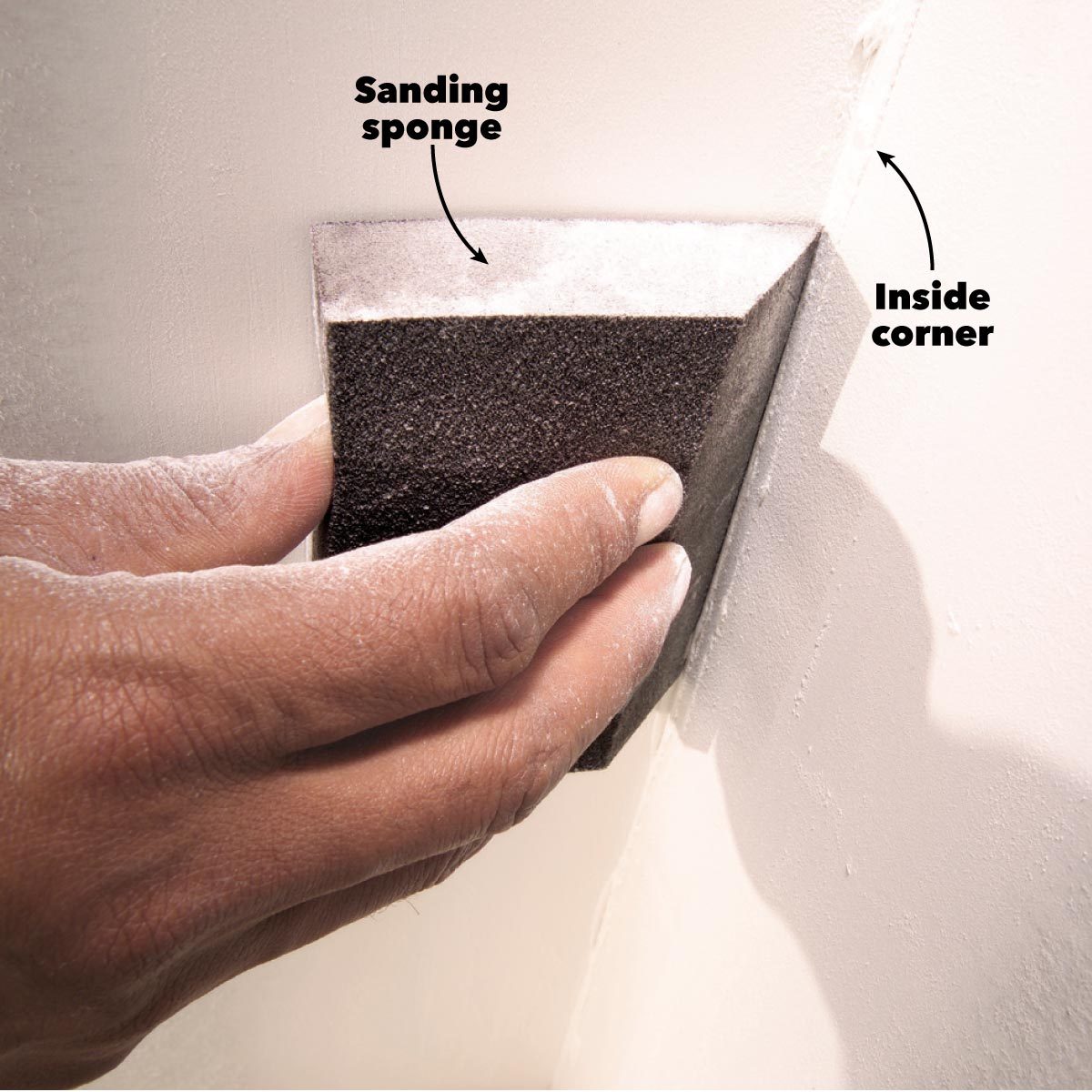 A hand holds a sanding sponge against an inside wall corner, smoothing the surface. The background shows a light-colored, textured wall.