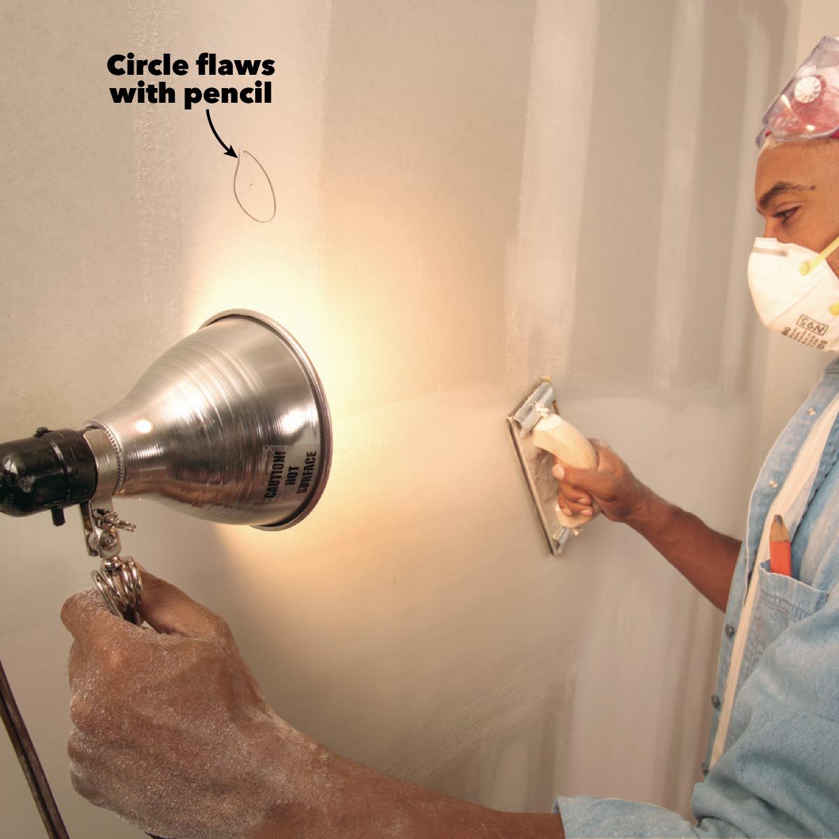 A worker is smoothing a wall with a sanding tool while illuminating flaws with a lamp and marking them with a pencil.