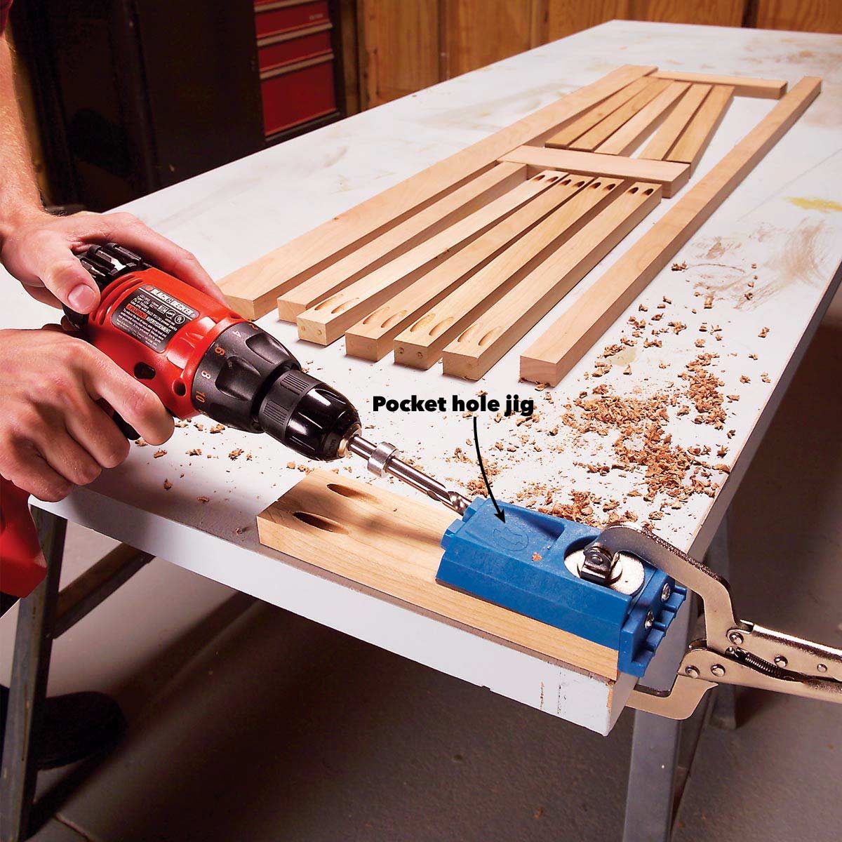 A person drills into wood using a power drill, guiding it with a blue pocket hole jig on a workbench surrounded by wooden strips and sawdust.