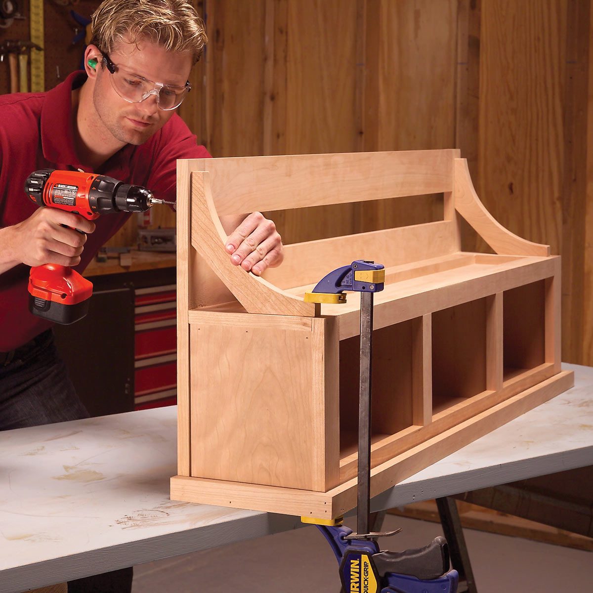 A man uses a drill to assemble a wooden bench on a table, supported by clamps, in a workshop with wooden walls and tools visible in the background.