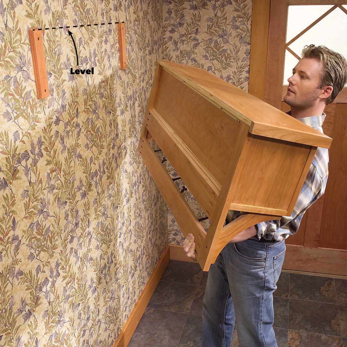 A man lifts a wooden object towards the wall, aligning it with two mounting brackets amid floral wallpaper and a wooden door.