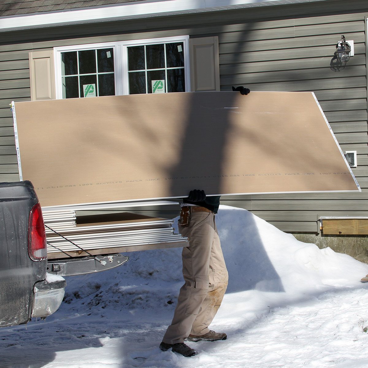 A person dressed in beige pants and black gloves carries a large sheet of material from a truck, surrounded by snow and a gray house.